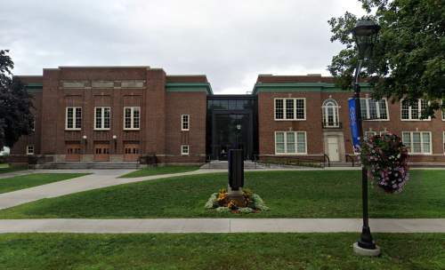 A brick building with large windows and a modern addition, surrounded by green grass and flower planters.